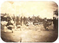 An old photograph showing a procession passing between lines of soldiers with tents in the background