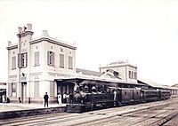 Photograph of a steam locomotive pulling passenger cars and sitting at the platform outside a large station building