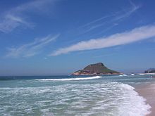 A landscape photograph taken along the shoreline of the Atlantic Ocean on a sunny day. Hills are visible in the background.