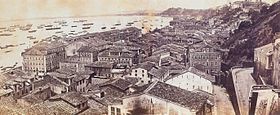 Photograph looking down a hillside overlooking the buildings of a town with a harbor beyond which is crowded with various vessels